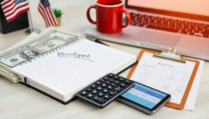 A desk with a notebook labeled "Budget," a calculator, a smartphone, a clipboard with financial documents, US dollar bills—perfect for tracking expenses or planning to avoid payday loans—plus a red mug, laptop, and two small American flags.