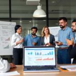 A group of six people stands and talks in an office meeting room, with charts and graphs on whiteboards. A laptop displaying Payday Loan Insights and data is open on the table in the foreground.
