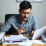 A man wearing glasses sits at a desk covered with papers, holding a magnifying glass and examining a document. He appears focused and serious, surrounded by stacks of paperwork and a calculator, as he investigates potential payday loan fraud.