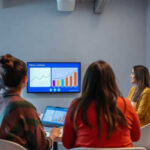 Four coworkers meet in an office to review fintech charts on a wall-mounted screen and a laptop.